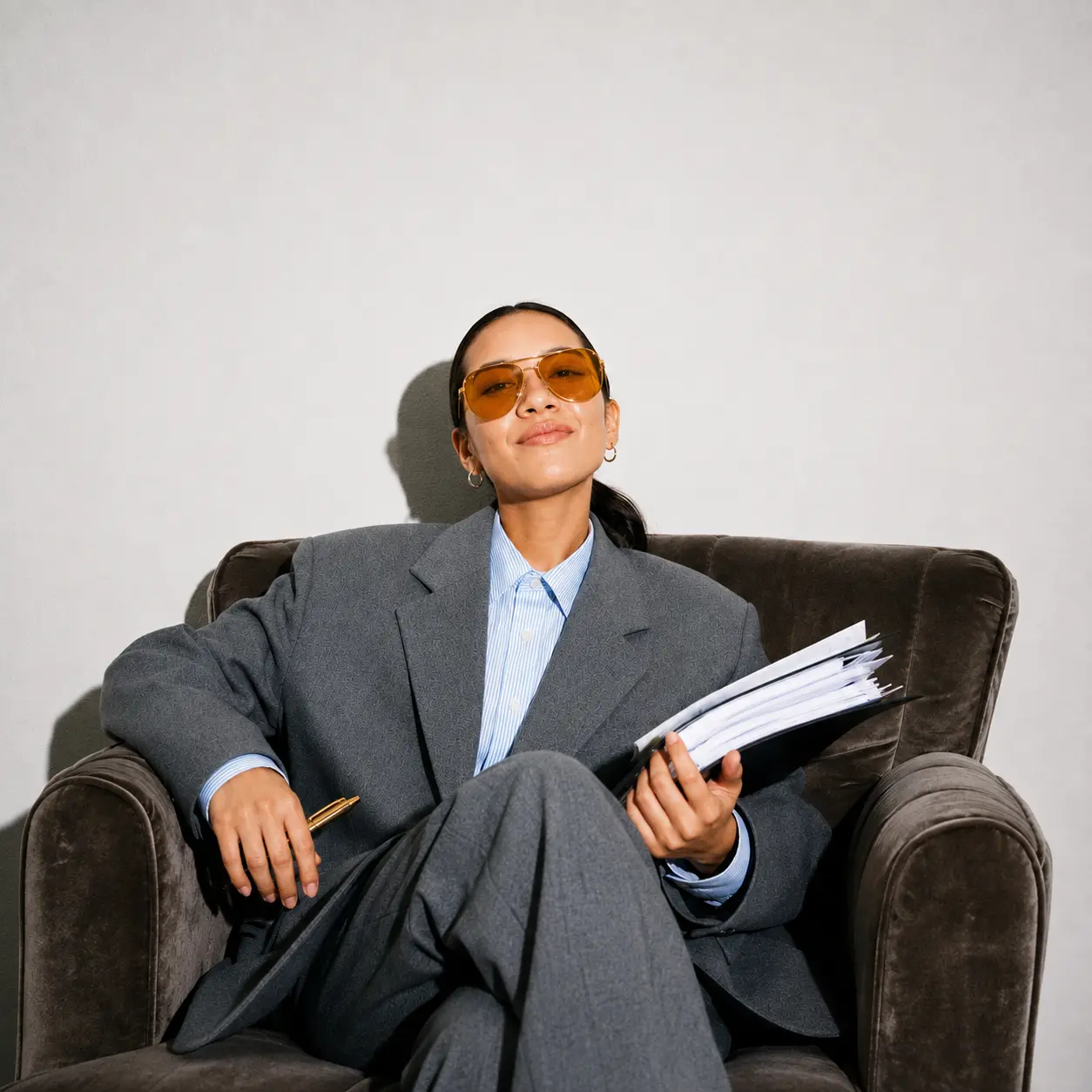 Eine Person in einem übergroßen grauen Anzug und einer orangefarbenen Sonnenbrille sitzt selbstbewusst in einem braunen Sessel, hält einen Stapel Papiere und einen Stift in der Hand, vor einem Hintergrund in neutralen Farbtönen.