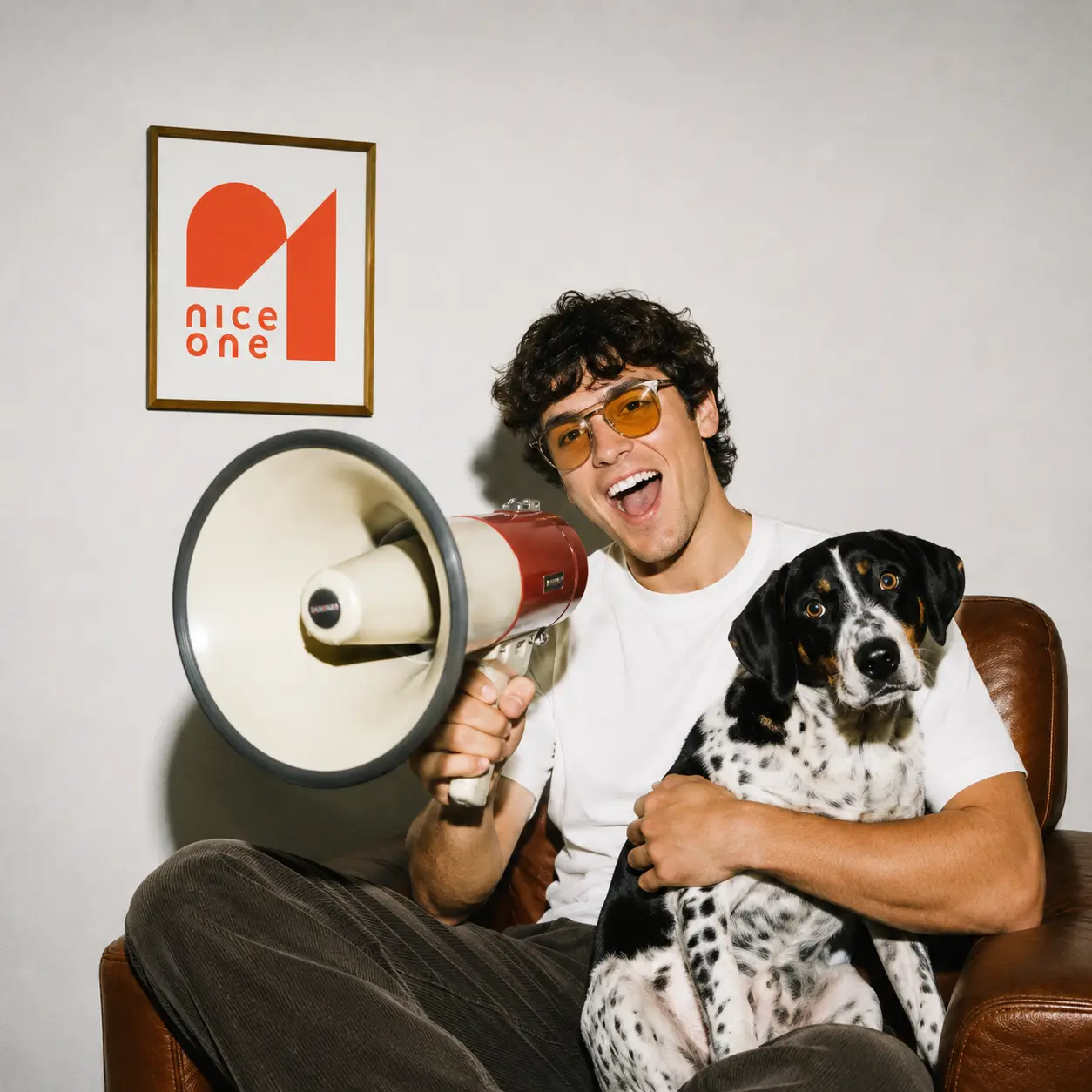 Ein lächelnder Mann mit Sonnenbrille hält ein Megafon in der Hand und sitzt zusammen mit einem schwarz-weißen Hund auf einem braunen Stuhl. An der Wand hinter ihnen hängt ein gerahmtes „Nice One“-Poster.