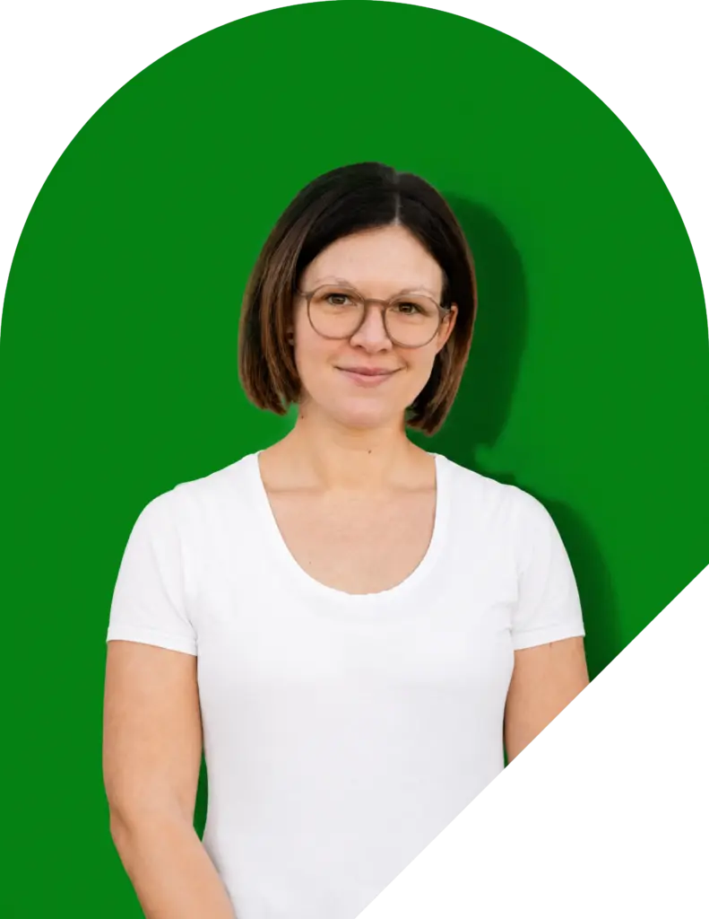 Eine Person mit kurzen braunen Haaren und Brille lächelt in die Kamera. Sie trägt ein weißes T-Shirt und steht vor einem hellgrünen Hintergrund, auf dem ein weißer Bogen zu sehen ist.